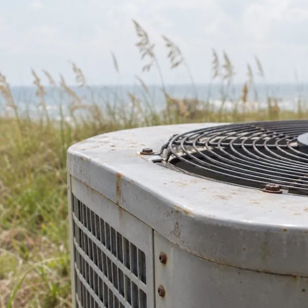 Weathered outdoor AC unit with rust spots, with beach dunes and the ocean behind it