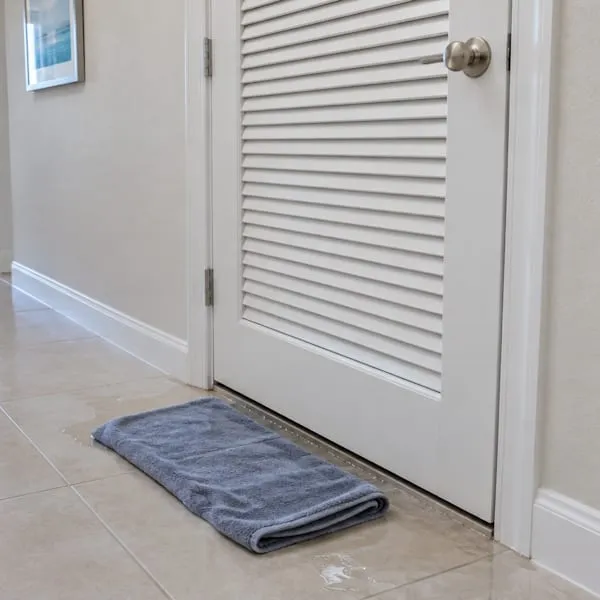 Folded towel soaking up water on a tile floor in front of a louvered air handler closet door